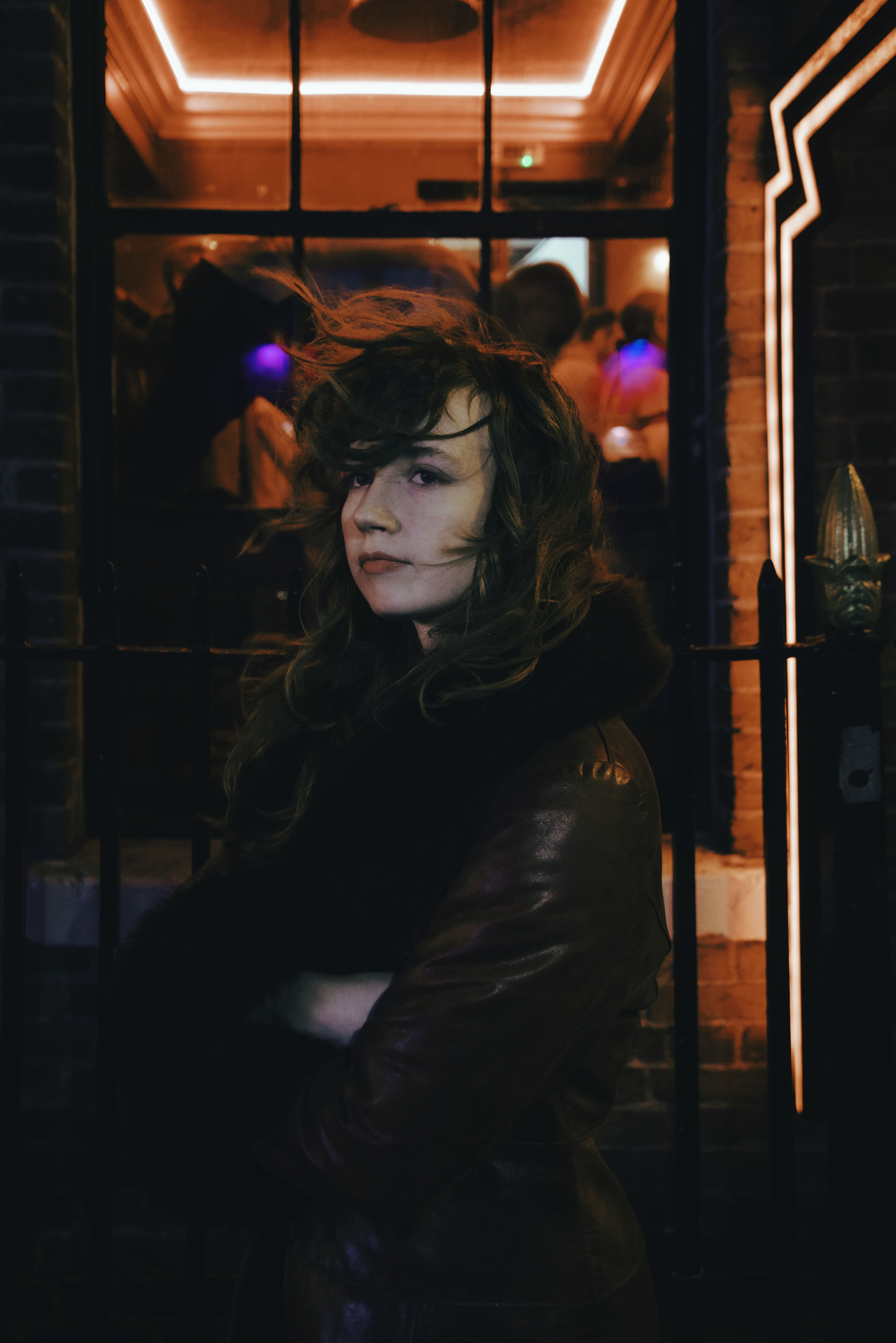 a woman with dark hair stands in a black coat outside of a building. the light through the window glows red