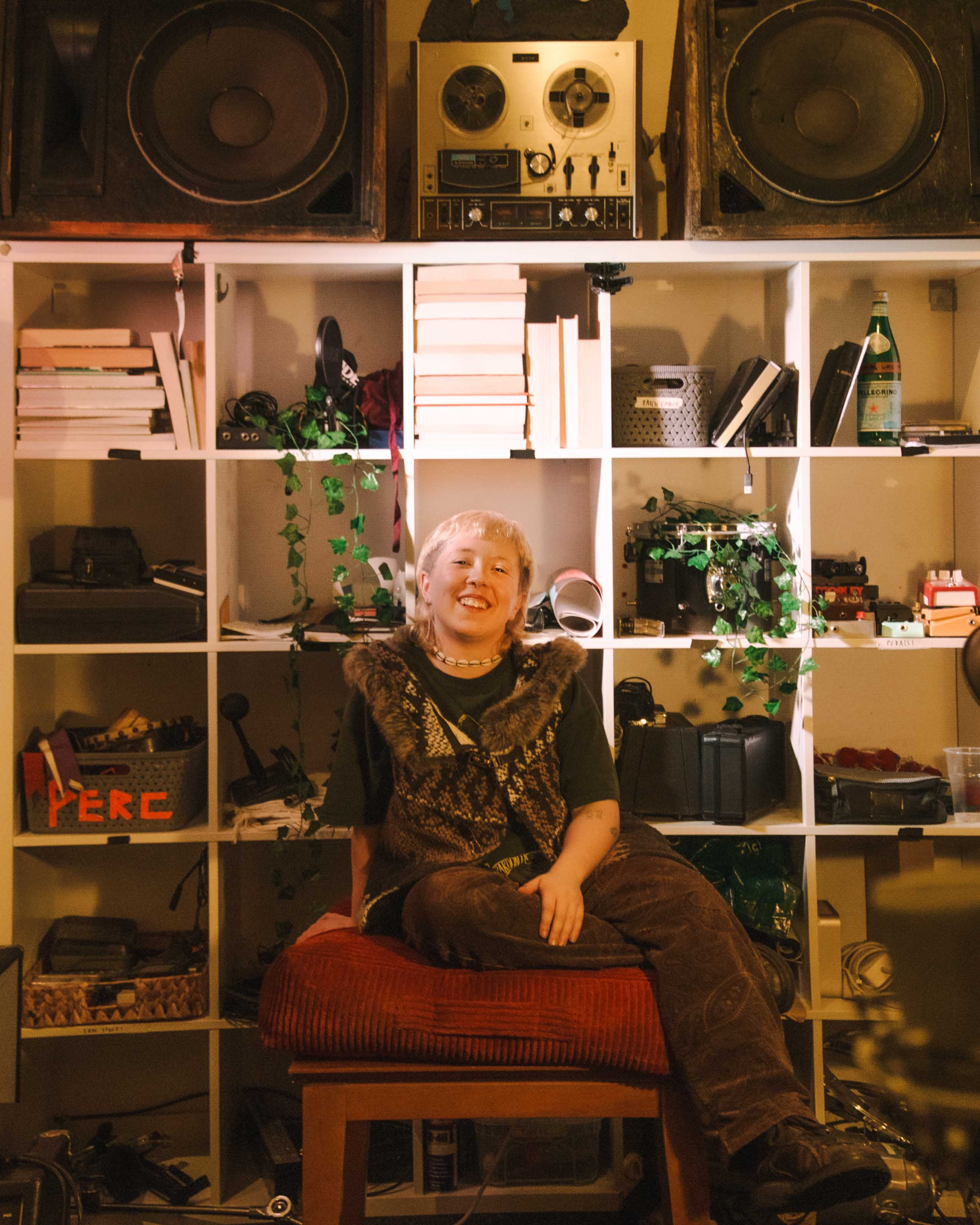 a person with blonde hair sits on a red cushion on a wooden stool and smles