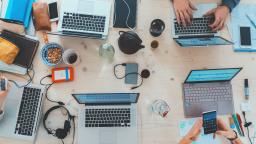 A birds eye view of a working space with hands typing on laptops on a desk, surrounded by notebooks, hard drives, and beverages.