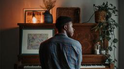 person with their back facing the camera, sitting at a piano, their head turned to look out the window