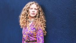 woman with long blonde curly hair in pink shirt against navy blue background