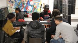 Group of people gathered around a piano, keyboard and guitar