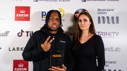 two people standing in front of white board with logos, one holding up award and holding up two fingers peace sign