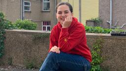 young person sitting outside with houses behind them, wearing blue jeans and a red hoodie