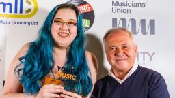 Young woman with long blue hair holding award, man wearing jumper next to her