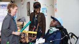 Woman standing and holding instruments, next to another woman smiling and a boy in a wheelchair wearing a cap