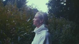 girl wearing white jumper with hair in a bun from a side profile, trees and green plants behind her