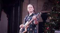 young woman with brown hair in pony tail playing a bass guitar. Christmas tree visible in right side of photo.