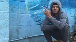 young person crouching down by a brick wall painted with blue graffiti