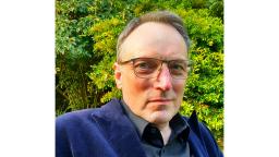 man with short hair and glasses cropped from shoulders up, green bushes in the background