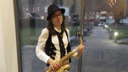 person wearing a white shirt and black waist coat, holding a saxaphone, standing in front of a glass door with houses and a street behind them.