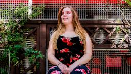 photo of a person sitting down, in the background there is a fenced wall with plants coming through it.