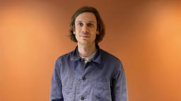 person wearing a navy blue shirt visible from the waist up. Standing against an orange lit background.