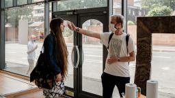 two young people standing inside a building next to hand sanitisers. one is giving the other a temperature check.