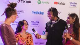 two presenters interviewing two artists in front of a step and repeat board