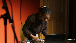 Musician bending over decks and a laptop, wearing headphones. There is the shadow of a mic stand in the foreground and a red wall in background.