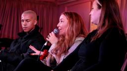 three people sit in a row. two are listening and one is holding a microphone and speaking as part of a panel.