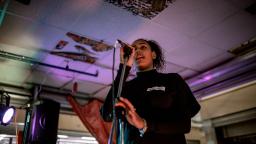 a young woman speaks or sings into a microphone on a stand. she wears a long sleeve black t shirt