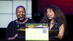 two women laugh on stage in front of a lecturn