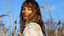 a woman with wavy brown hair and a fringe looks at the camera. she is in a wheat field