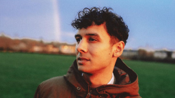 a man with dark curly hair looks to the side in a park. he is wearing a hooded jacket