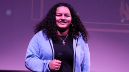 a person with dark curls holds a microphone and wears a blue hoodie unzipped over a black top