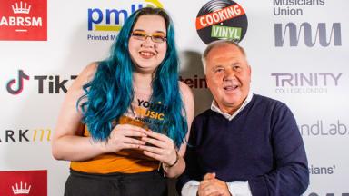 two people standing in front of background with logos printed on it. one holds an award