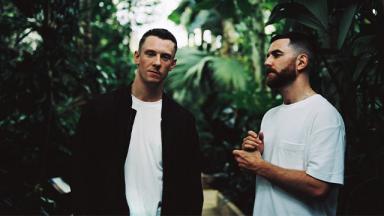 two people in white tshirts with green jungle trees behind them