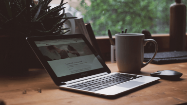 open macbook and mug on a wooden desk in front of a window