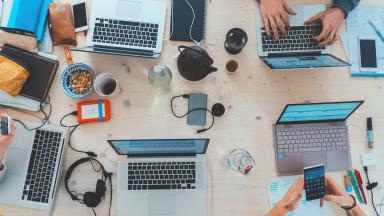 A birds eye view of a working space with hands typing on laptops on a desk, surrounded by notebooks, hard drives, and beverages.