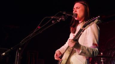 Person with shoulder length brown hair and wearing all white singing on stage and playing guitar.