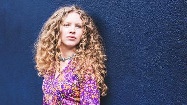 woman with long blonde curly hair in pink shirt against navy blue background