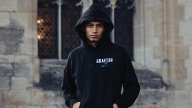 person wearing black hoody with hands in pockets. Brick building with large window in the background.