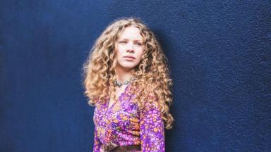 young person from waist up wearing a pink blouse, long curly blonde hair, looking away from camera, against a blue background