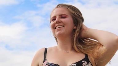 person visible from chest up, smiling into the distance with one hand running through their hair. A blue sky and clouds visible in the background.