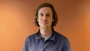 person from chest up wearing a navy blue shirt, standing against an orange lit background