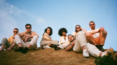 seven people sitting on grassy hill with blue sky in background.