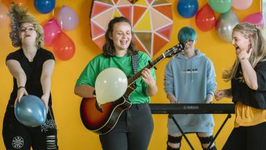 Group of four musicians in front of a yellow wall with balloons