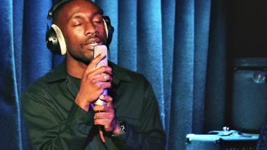 a man with a septum piercing sings passionately into a microphone. he is standing against a blue velvet curtain.