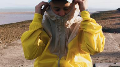 luke Rv sits on the beach, wearing a yellow jacket, fawn trousers and a blue cap.