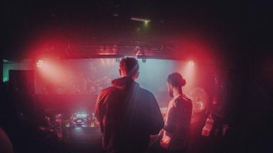 BICEP play a gig for Youth Music. The lighting is red and the duo are behind the DJ decks in front of a crowd below.