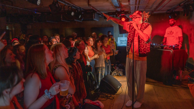 jordan stephens stands on stage and sings into a microphone. a crowd looks up at him as he is bathed in red lighting