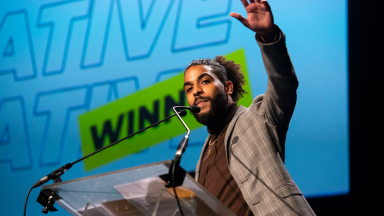 superlative raises his left arm on stage at the youth music awards. the word winner is on the screen behind him.