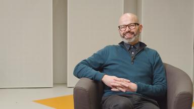 anthony hamer-hodges sits in a chair and smiles. he wears a blue jumper and glasses.