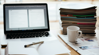 a laptop screen showing a word document, a stack of magazines and coffee cup