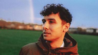 a man with dark curly hair looks to the side in a park. he is wearing a hooded jacket