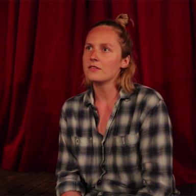 woman wearing black and white checkered shirt, sitting in front of a red curtain, looking away from the camera