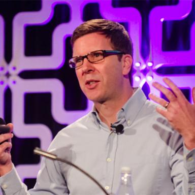 man wearing light blue shirt, gesturing with hands and speaking into microphone