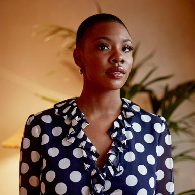 woman with shaved head wearing blue polkadot shirt, plant visible in background