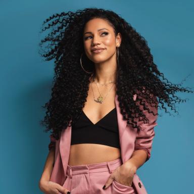 woman with black curly hair wears dusky pink blazer and short combo with black crop top as she looks to the right